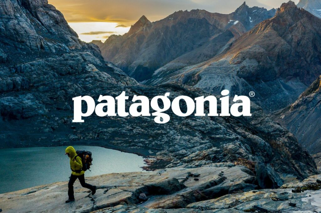 Hiker in yellow jacket with backpack walking on rocky terrain in front of mountain landscape, Patagonia logo in foreground.