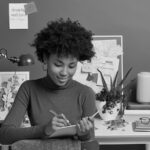 Woman writing in a notebook at a modern home office desk with a bulletin board in the background.