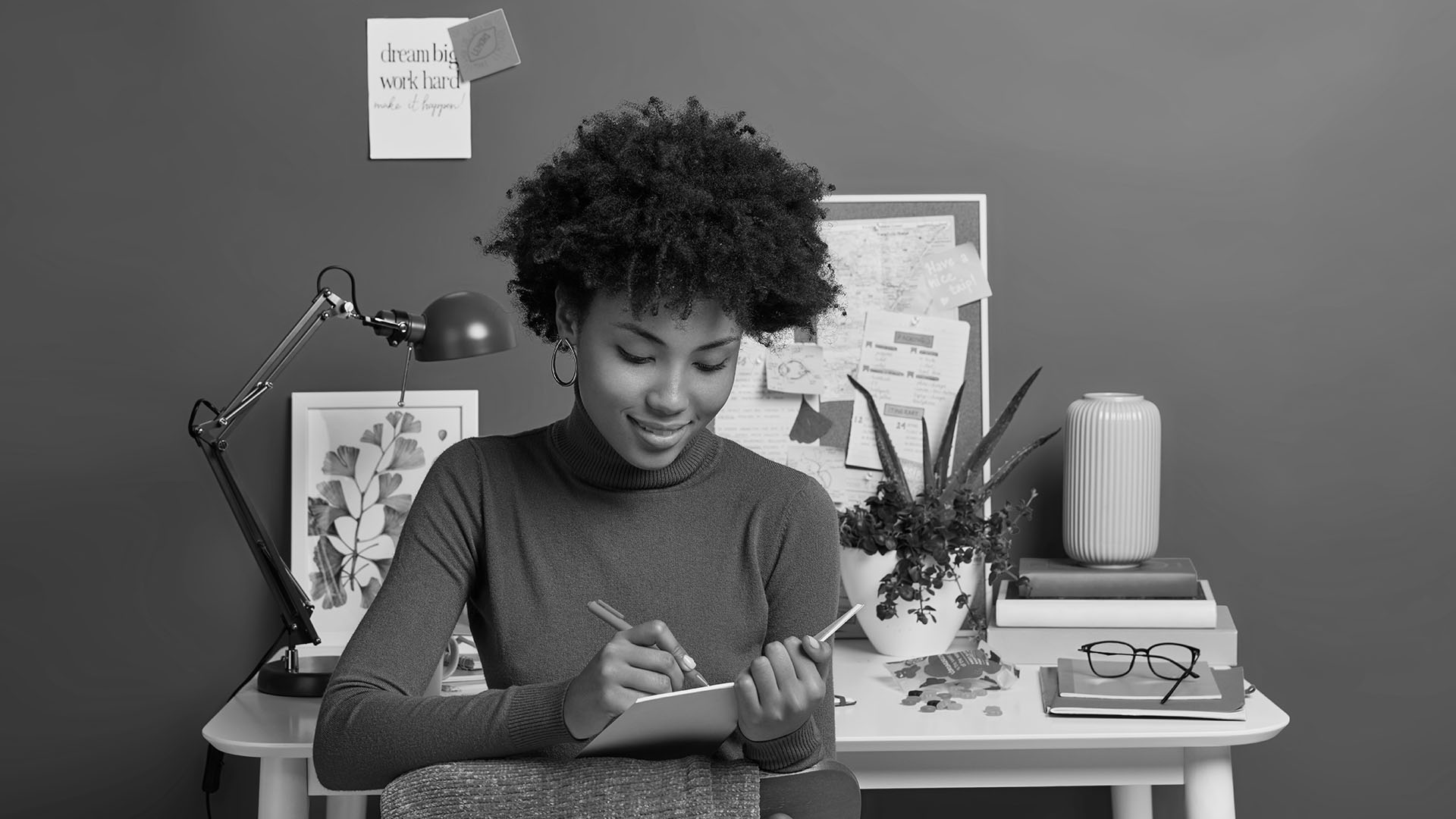 Woman writing in a notebook at a modern home office desk with a bulletin board in the background.