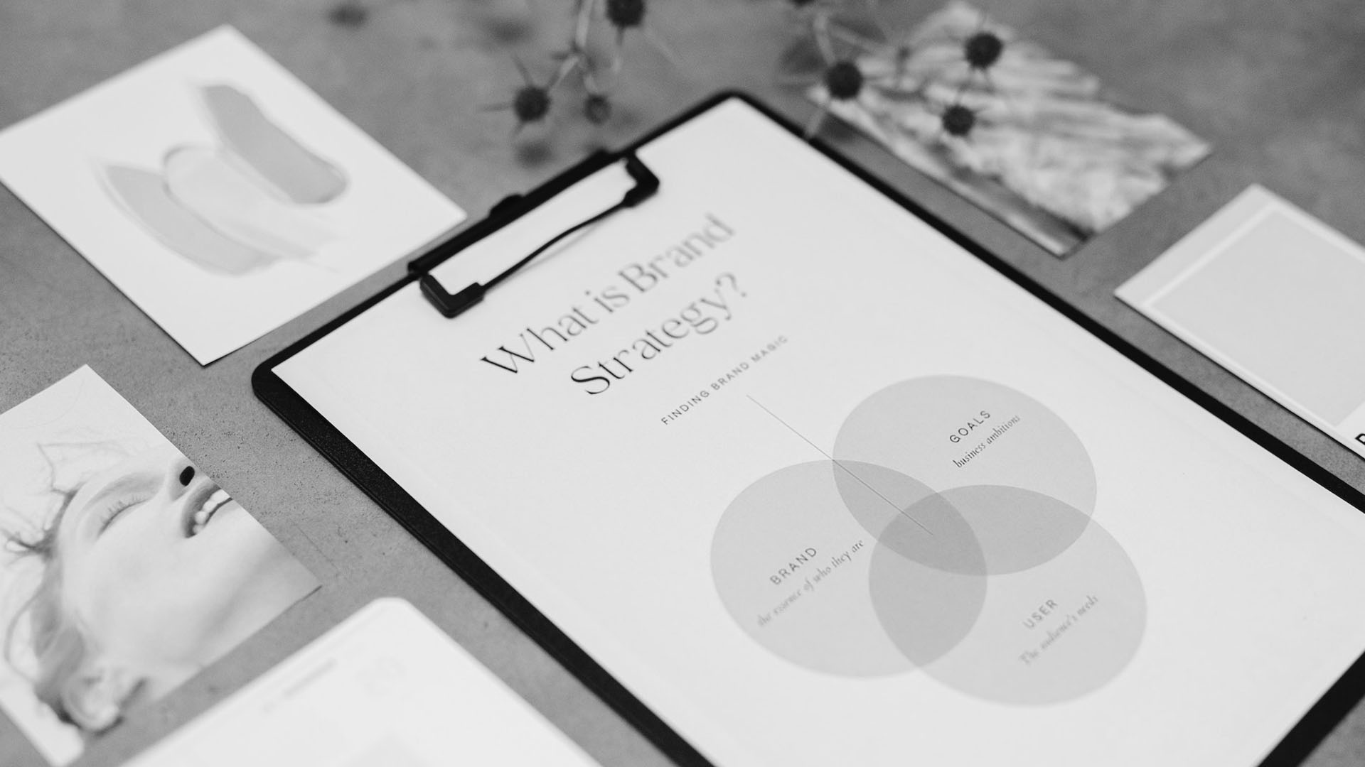 Black and white image of a brand strategy Venn diagram on a clipboard with design samples around it.