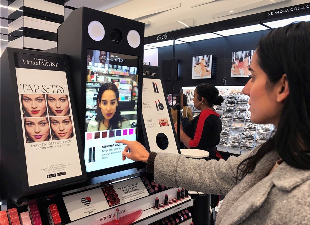 Woman using a Sephora Virtual Artist “Tap & Try” kiosk in a store; the screen shows her face with virtual lipstick colors and shade options.